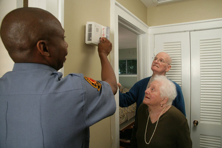 A fire safety official shows an older man and woman how to test a smoke alarm.