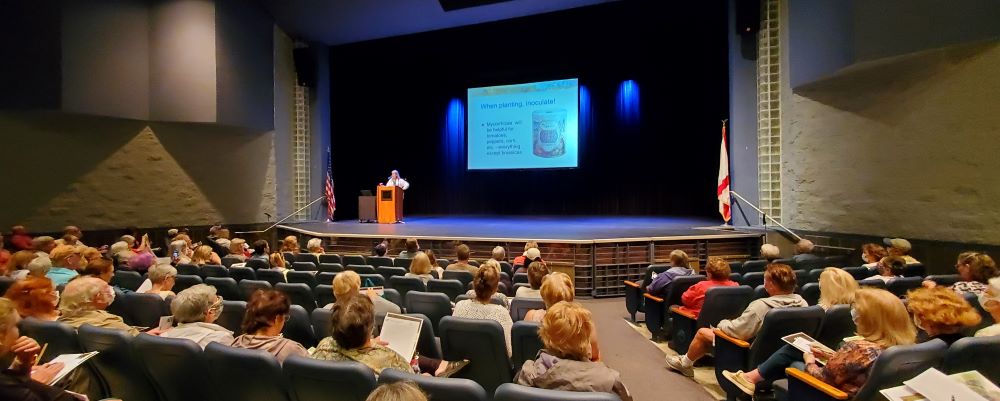 An audience in the Eau Gallie High School Auditorium watches a woman give a presentation on stage.