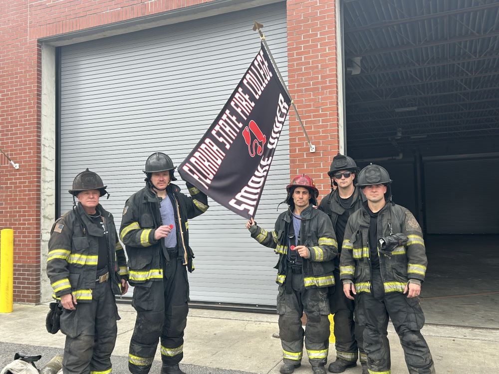 Payton Jones, Zac James, Tony Mauro, Patrick Milbocker and Steve White in full gear standing in front of the Florida State Fire College Smoke Diver flag.