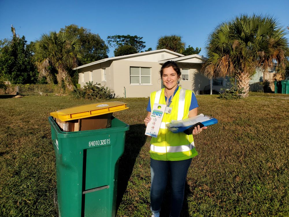 City worker in safety vest  standing next to a full recycling cart and holding an informational door hanger