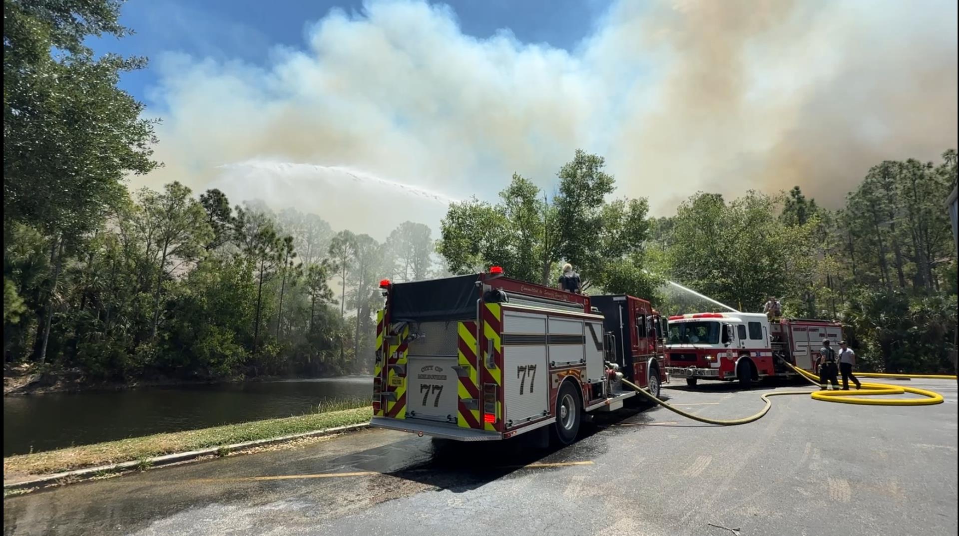 Firefighters shoot water from hoses off two fire engines into a wooded area with a great deal of smoke rising in the background.